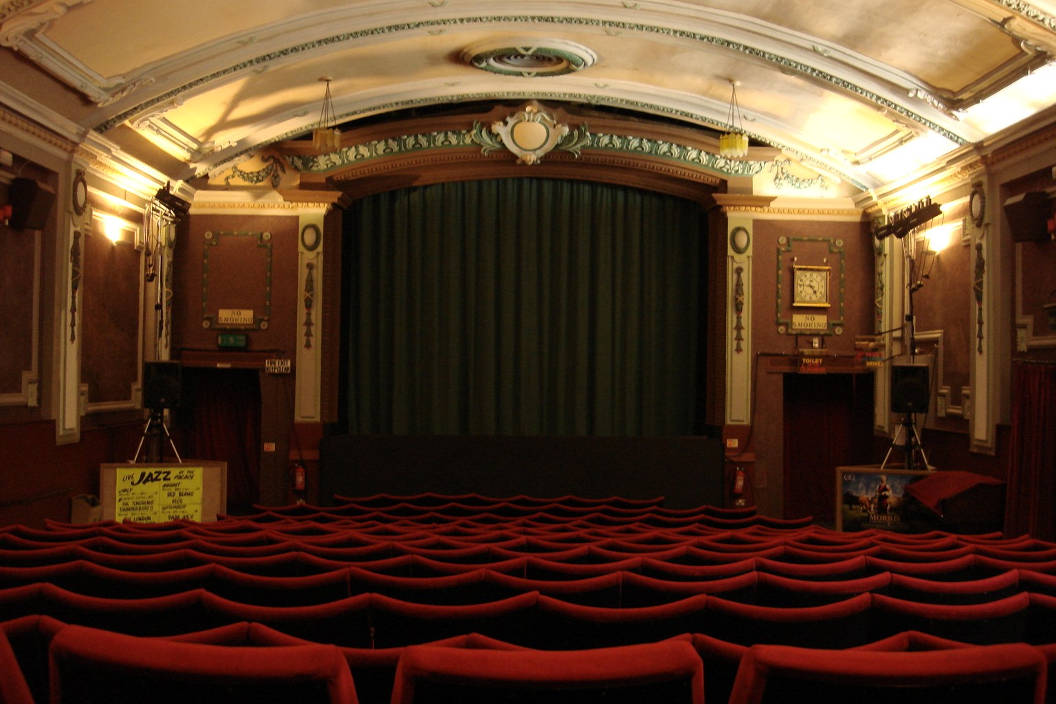 Interior of a small historical cinema seen from the back: empty rows of red velvet seats, a closed dark green stage curtain, ornate Edwardian plasterwork, dim warm light.