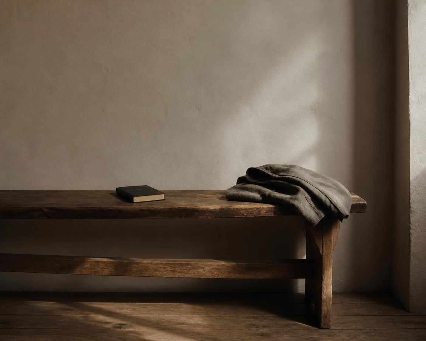 A worn wooden bench against a plain wall in late afternoon light, with a small closed book and a folded jacket resting on it.