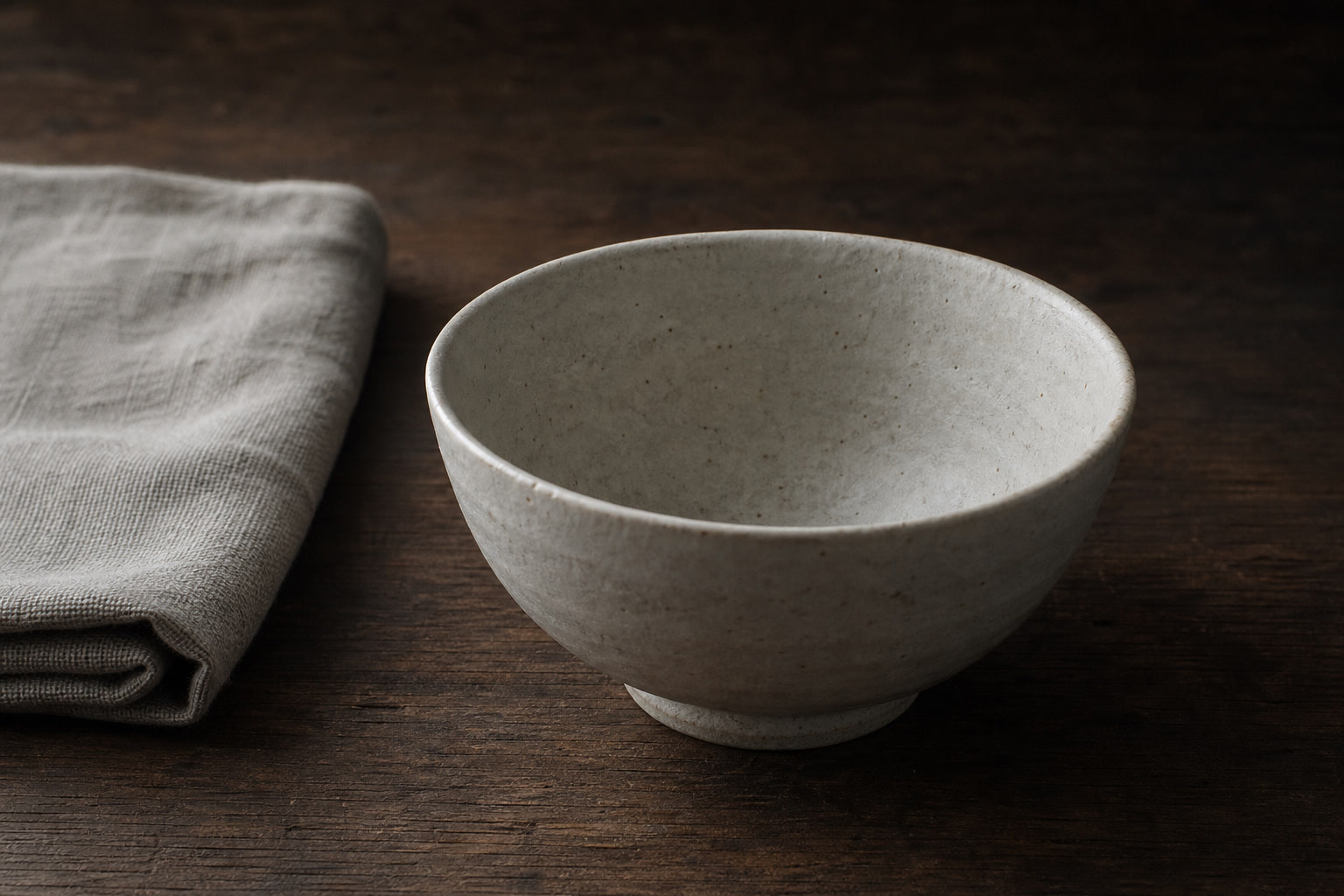 A plain unglazed Korean-style rice bowl set on a dark wooden table in soft northern light, with a folded coarse linen cloth to one side.