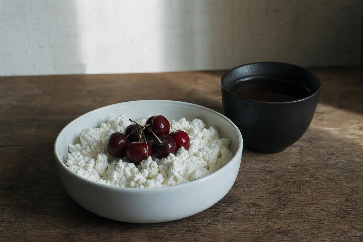 A quiet breakfast still life: a bowl of ricotta and cherries beside a dark cup of black tea on a wooden table in morning light.