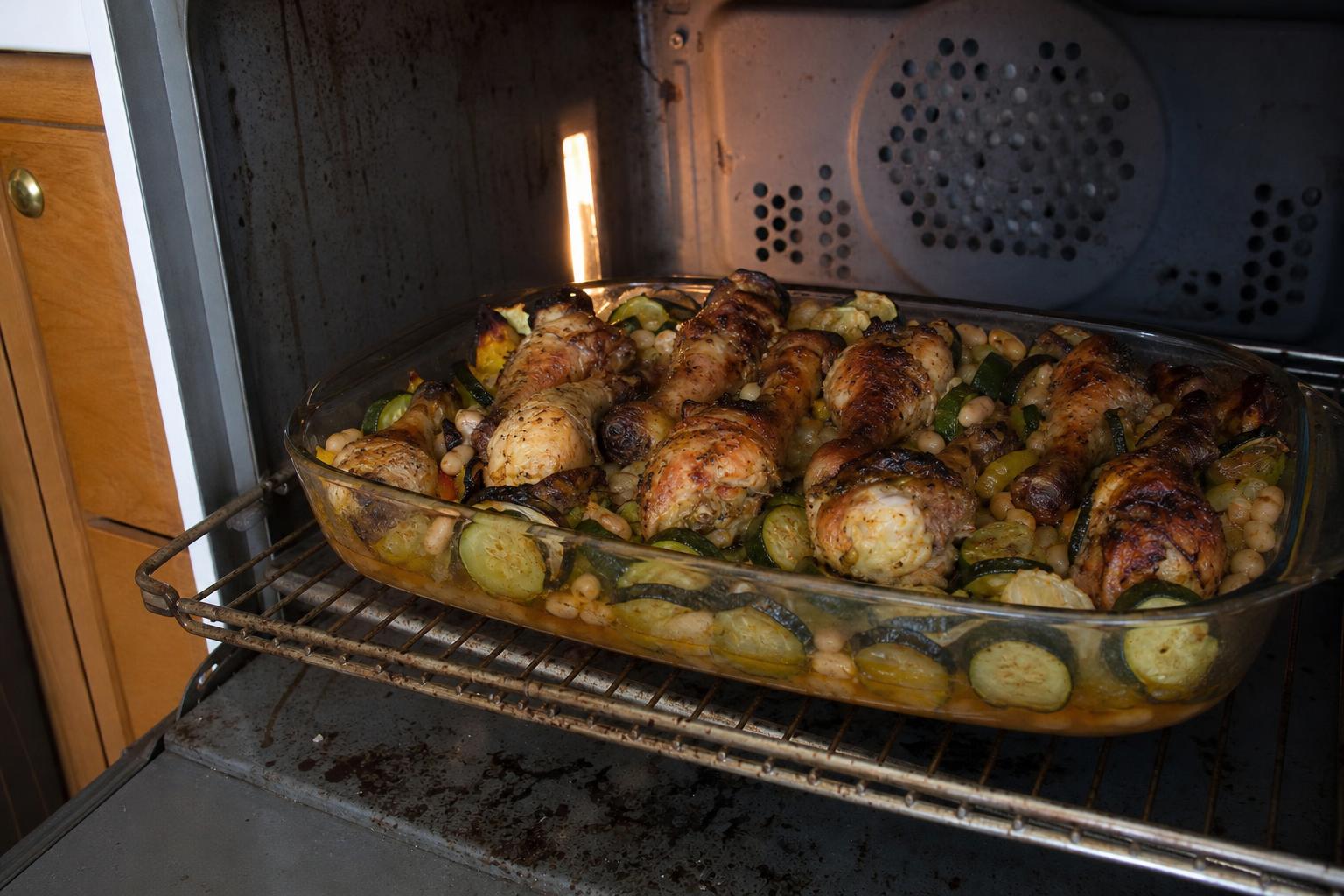 Restrained editorial image of browned chicken drumsticks over white beans and sliced vegetables in a glass roasting dish at the oven door.