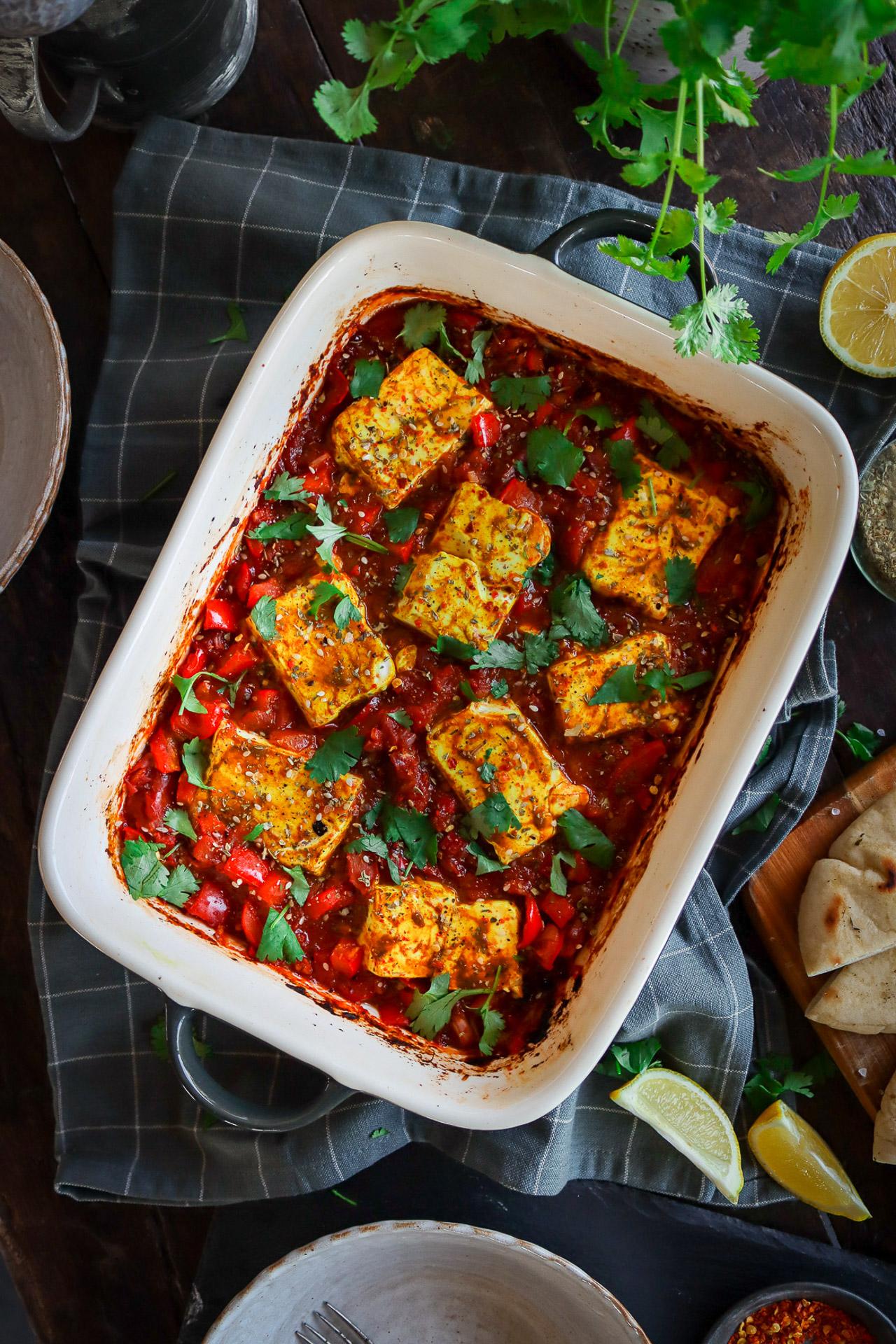 Ofen-Tofu-Shakshuka in einer schweren Pfanne: rote Paprika-Tomaten-Sauce, gelb gewürzte Tofuscheiben, frische Kräuter.