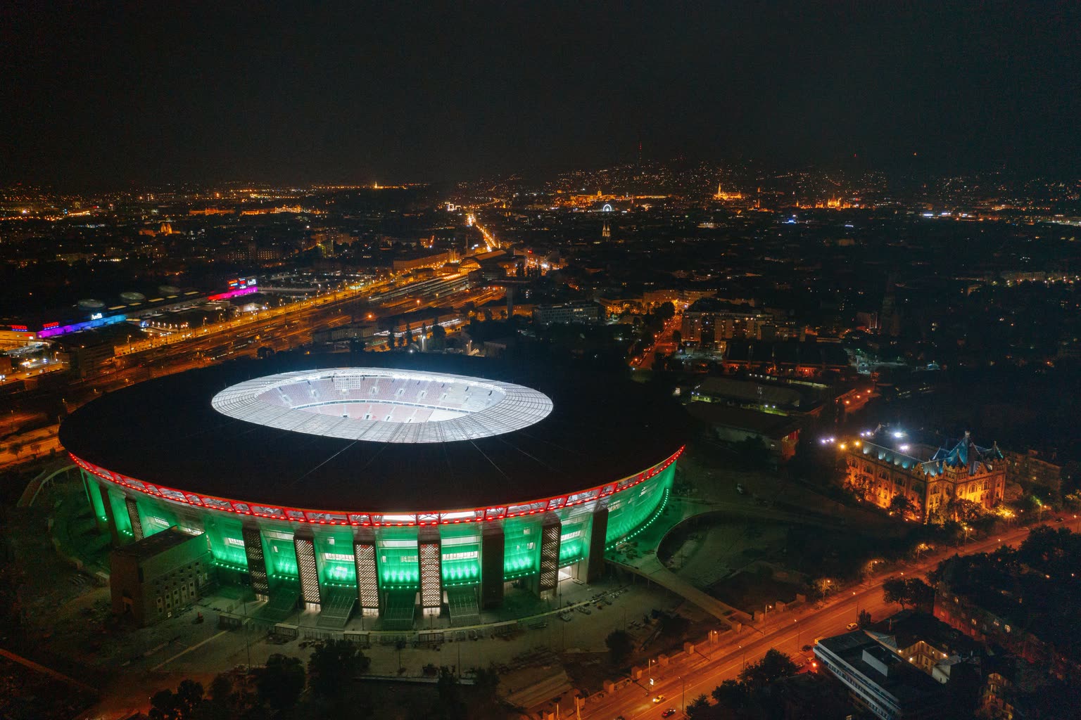 Aerial night photograph of an empty Puskás Aréna in Budapest, the bowl lit green and red, with the dark city stretching out behind it to a dim horizon.