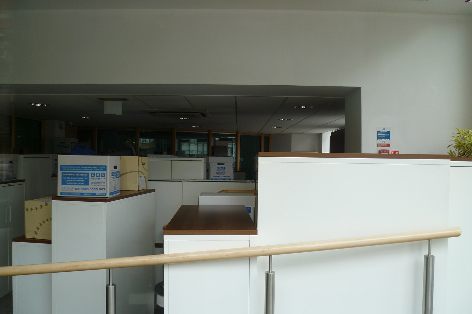 Empty interior of a closed Laiki Bank branch in North London after closure, with white teller counters, moving boxes stacked on the desks, and grey daylight from above.