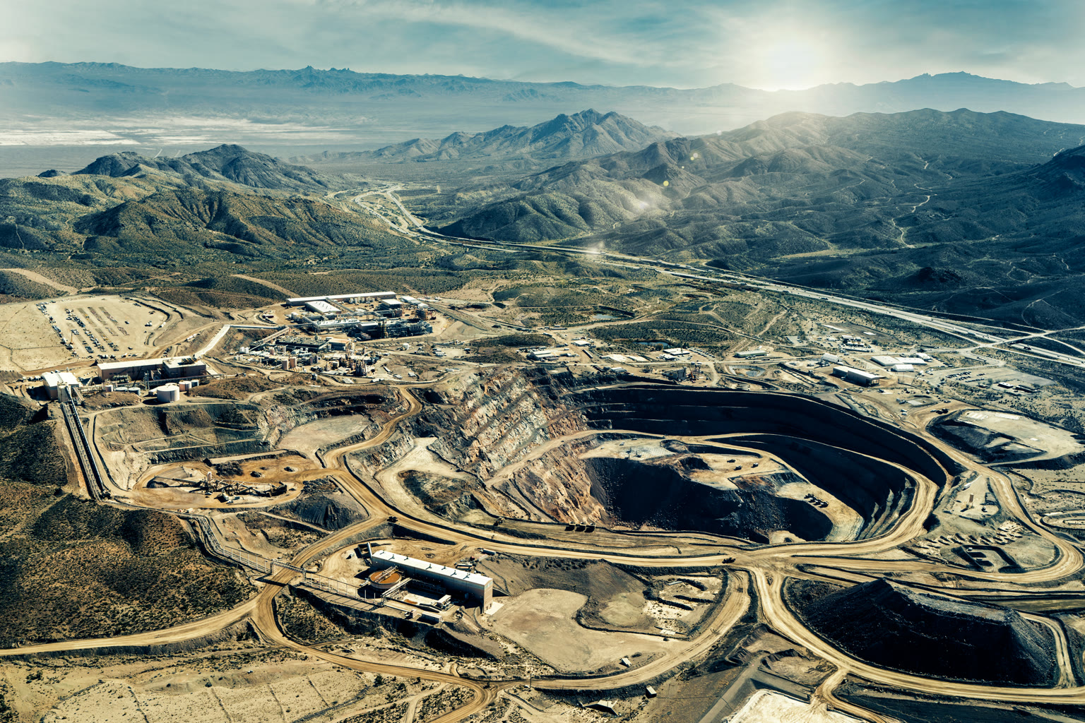Aerial photograph of the Mountain Pass rare earth mine and processing facility in the Mojave Desert: a deep open pit dominates the frame, with the modest cluster of processing buildings to one side and an empty dry landscape stretching to distant mountains.