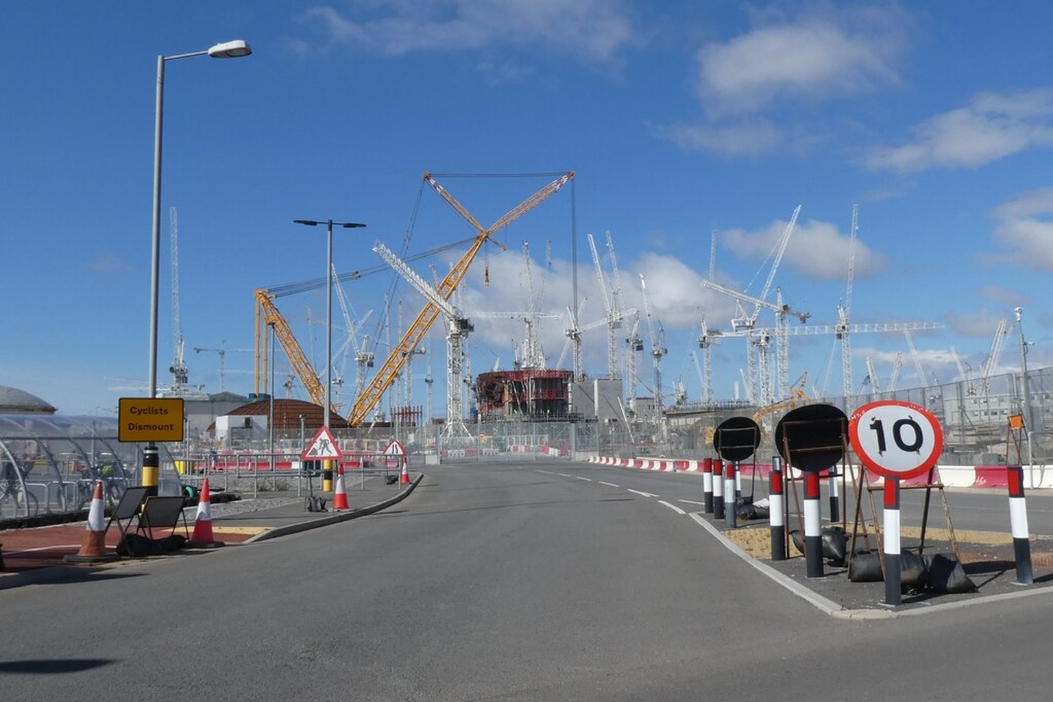 A forest of construction cranes at Hinkley Point C, with cones, speed-limit signs and a yellow Cyclists Dismount notice across the foreground road.