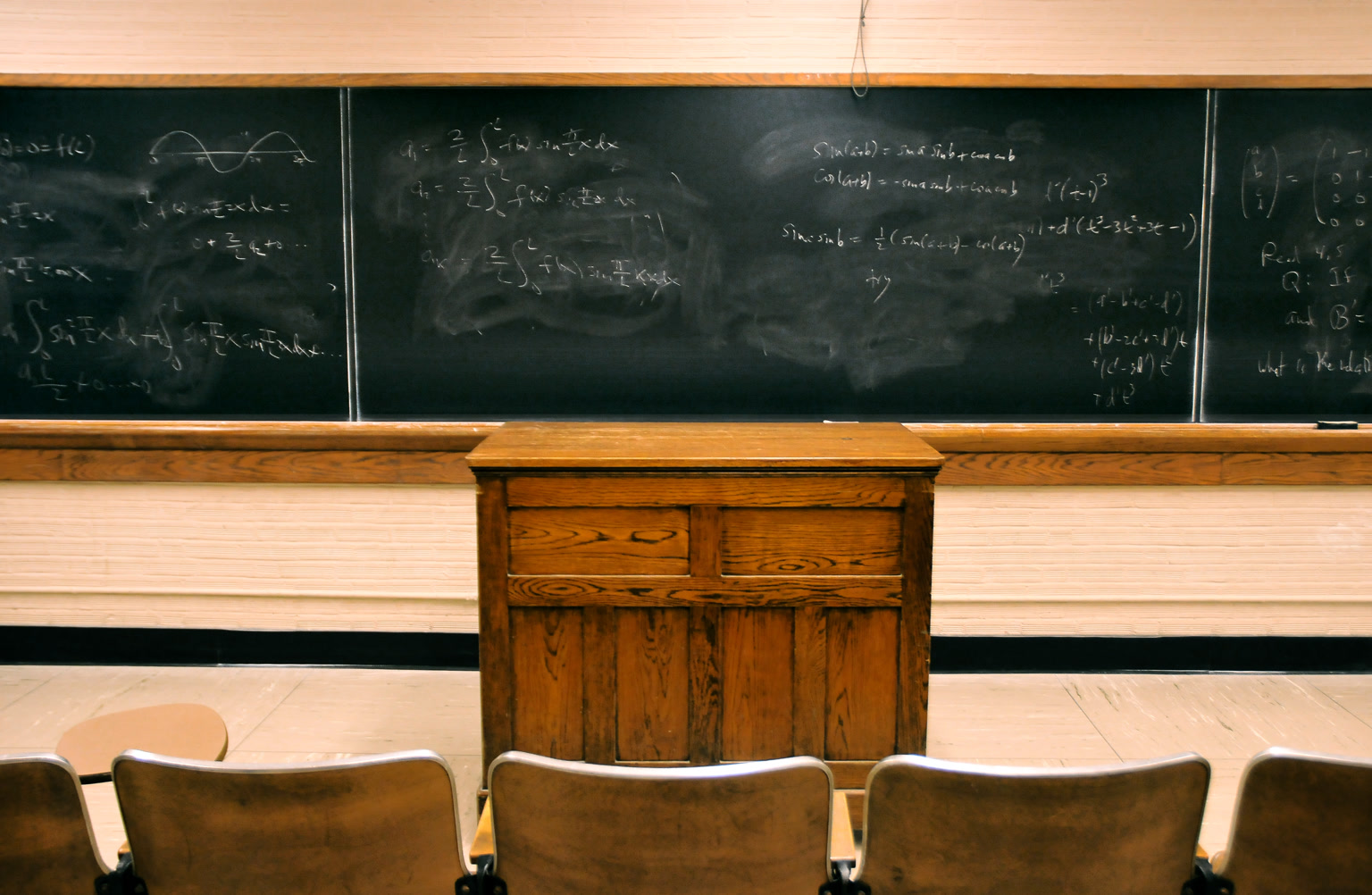 A classroom blackboard dense with chalked mathematical formulas, diagrams, and crossed-out work, suggesting a room where thought must be worked through rather than instantly fetched.